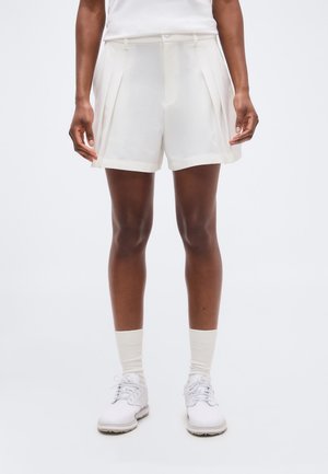 Osoba oblečená v bílých plisovaných šortkách, bílých ponožkách a bílých sportovních botách na jednobarevném pozadí.