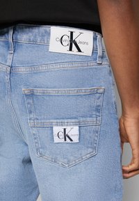 Ljust blå jeans med Calvin Klein Jeans-lappar på baklinning och ficka, bärs av en person med mörk hy och en svart topp.