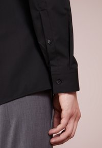 Rechter Arm trägt ein schwarzes langärmliges Hemd mit geknöpftem Manschettenrand, ruht neben grauer Hose vor neutralem Hintergrund.