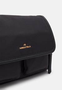 Borsa nera in nylon con patta, caratterizzata da un logo dorato e una texture liscia. Include due tasche frontali e dettagli di cuciture a contrasto.