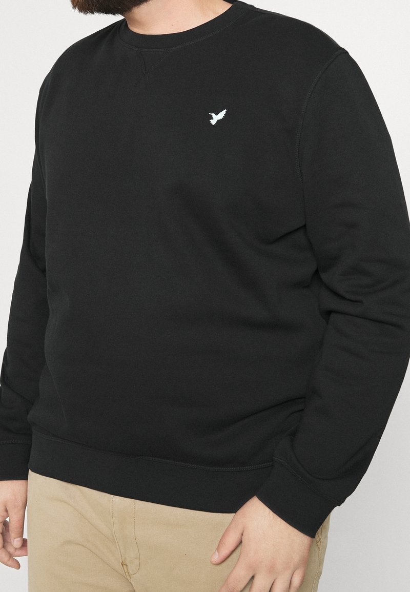 Homme portant un sweat-shirt noir à manches longues avec un petit logo d'oiseau blanc sur la poitrine et un pantalon beige.