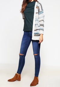 Tjockstickad cardigan i vitt med flerfärgade ränder, parat med en mörkgrön ribbad topp och blå skinny jeans med trasiga detaljer, samt tanfärgade ankelboots.