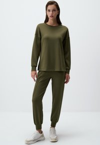 Completo felpa e jogger verde oliva, caratterizzato da una vestibilità comoda, maniche lunghe, polsini a costine e texture morbida. Abbinato a sneakers di colore chiaro.