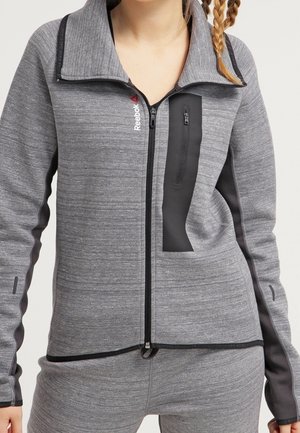 Giacca sportiva grigia con un alto colletto, dotata di una tasca con zip nera, tessuto testurizzato e pannelli laterali neri a contrasto. Logo Reebok visibile.