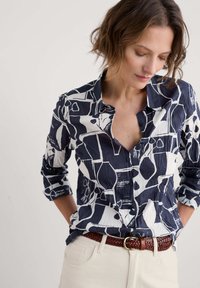 Jūras zilās un baltās krāsas rakstainā krekls ar apkakli, pogām un sarullētām piedurknēm. Valkāts ar pīta brūna josta un gaismas krāsas biksēm.