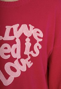 Pull en tricot rouge avec un col rond, orné d'une broderie en rose clair qui indique "L'amour est l'amour" dans différentes polices et tailles.