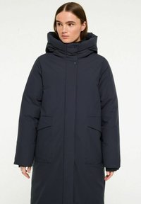 Lång mörk marinblå puffjacka med hög krage, dold dragkedja, stora framfickor och strukturerad yta. Har även huva och knapprich.