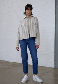 Striped, cropped white jacket with two front pockets, paired with blue jeans and white sneakers with pink accents. Smooth textures overall.