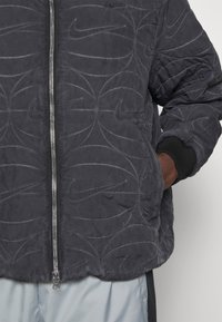 Giacca trapuntata grigio scuro con chiusura zip, caratterizzata da loghi Nike in rilievo disposti in motivi vorticosi. Maniche a costine e vestibilità comoda.