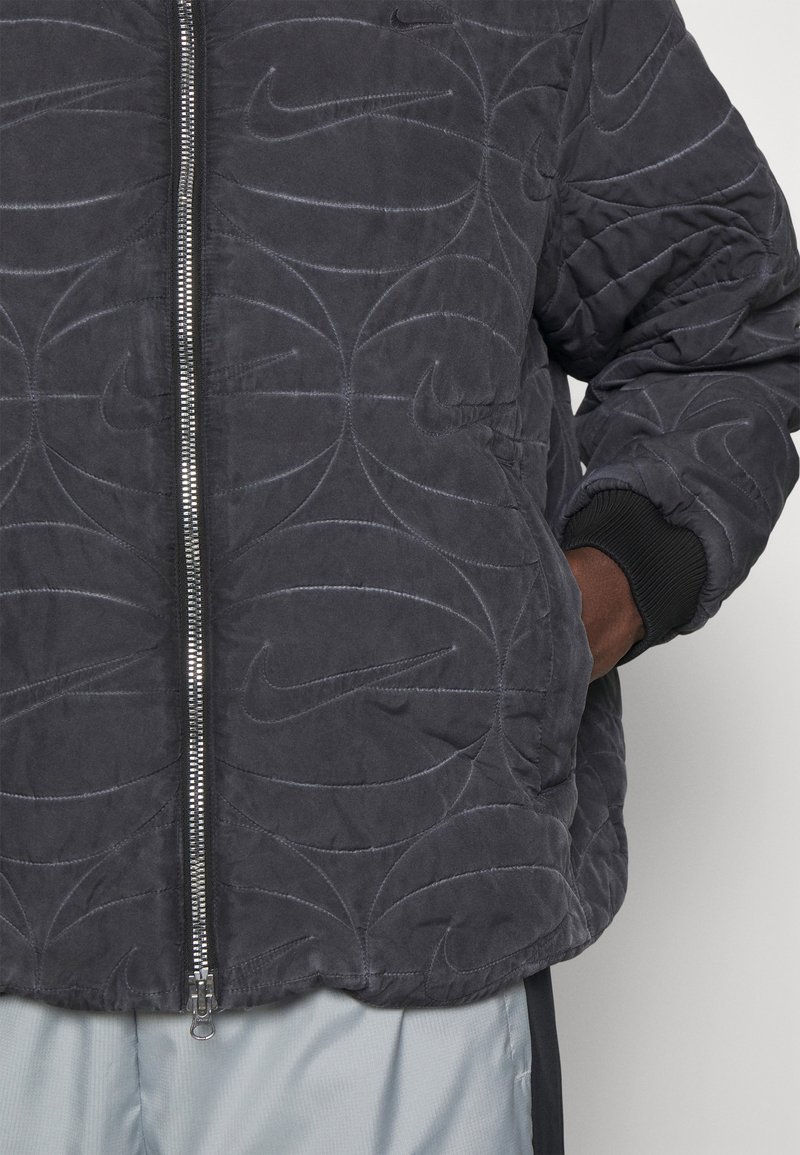 Giacca trapuntata grigio scuro con chiusura zip, caratterizzata da loghi Nike in rilievo disposti in motivi vorticosi. Maniche a costine e vestibilità comoda.