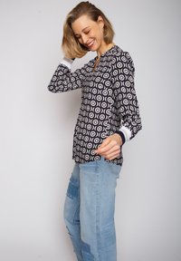 Schwarz-weiße gemusterte Bluse mit kreisförmigen Designs, langen Ärmeln und V-Ausschnitt, kombiniert mit hellblauen, ausgewaschenen Jeans.
