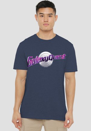 Marineblaue Baumwoll-T-Shirt mit lila "Galaxy Quest"-Schriftzug und einem kreisförmigen silbernen Grafik. Kurze Ärmel, standardmäßiger Rundhalsausschnitt.