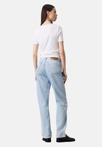 Jeans de mezclilla azul claro con un corte relajado, que cuentan con bolsillos traseros clásicos y un parche de cuero marrón en la cintura. El modelo lleva una blusa blanca.