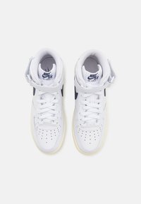 Ett par vita Nike Air Force 1 högskor med kardborreband och svarta swoosh-logotyper, sedda ovanifrån på en enkel bakgrund.