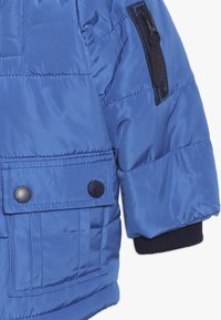 Sininen puffatakki, jossa on pehmeä ja kiiltävä pinta, vetoketjullinen tasku hihassa ja suuret napit etutaskussa.