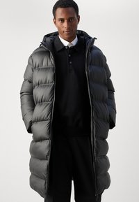 Lange pufferjas in matte zwart, met een capuchon, ritsluiting en gewatteerde textuur met grote delen gevuld met dons. Gekocht over een zwarte polo.