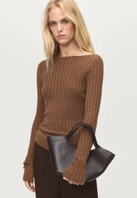 Brauner gerippter Pullover mit weitem Ausschnitt und langen Ärmeln. Schwarze Ledertasche mit geschwungener Form und kurzem Griff, die in der Hand gehalten wird.