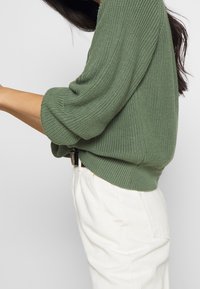 Pull en maille verte avec manches surdimensionnées, texture côtelée et un ourlet qui chevauche un pantalon blanc doté d'une poche latérale et d'une coupe décontractée.