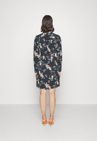 Abito floreale con maniche lunghe, base nera, presenta fiori rosa, verdi e bianchi. Include una cintura coordinata. Indossato con tacchi alti arancioni.