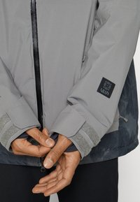 Veste grise imperméable avec une fermeture éclair noire, des poignets en velcro gris et une étiquette noire de marque sur la manche. Tissu texturé à deux couches.