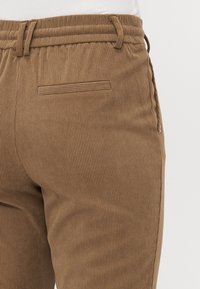 Brune kordfløjspants med en struktureret overflade, der har en snor i taljen, sidelommer og en ren, syet baglomme detalje.