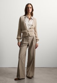 Cardigan corto beige a maglia con bottoni, sopra una camicia beige chiaro. Abbinato a pantaloni beige a gamba larga e scarpe marroni chiuse.