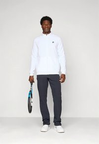 Chaqueta deportiva blanca con cremallera, logo negro en el pecho, combinada con pantalones deportivos gris oscuro. Sosteniendo una raqueta de tenis con detalles en azul.