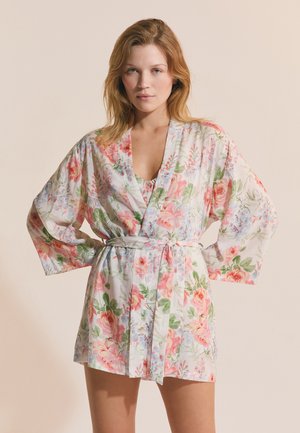 Frau trägt einen kurzen Bademantel mit Blumenmuster in Rosa und Grün, der in der Taille gebunden ist, steht mit den Händen in die Hüften gestützt.