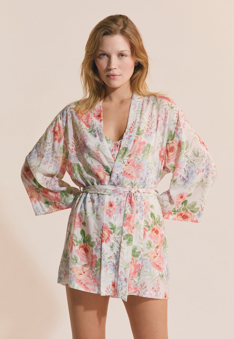 Frau trägt einen kurzen Bademantel mit Blumenmuster in Rosa und Grün, der in der Taille gebunden ist, steht mit den Händen in die Hüften gestützt.