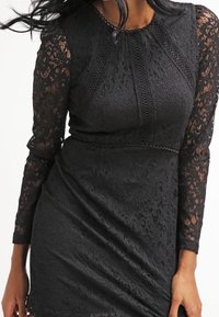 Svart spetklänning med långa ärmar, som har intrikata blommönster, en figurnära bodice och en slät kjol med en lätt rynka.