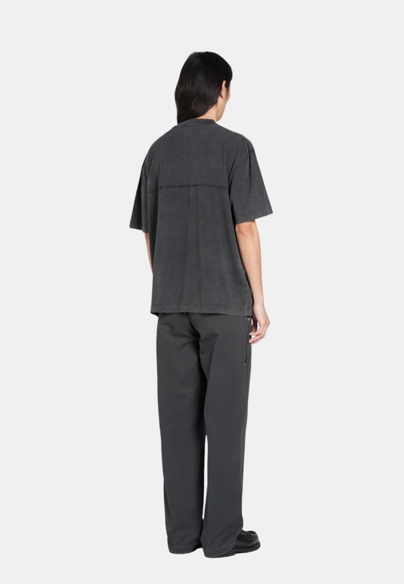 Personne aux longs cheveux noirs portant un t-shirt noir délavé ample et un pantalon noir droit, debout de dos sur fond blanc.