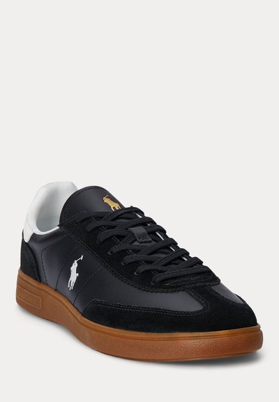 Zwarte lage sneaker met zwarte veters, wit logo aan de zijkant, gouden logo op de tong, witte hielstrip en bruine rubberen zool.