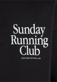 Felpa nera con caratteri bianchi audaci: "Sunday Running Club" e "ANOTHER COTTON LAB" stampati nella parte anteriore con dimensioni di carattere diverse.