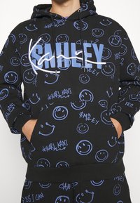 Osoba v černé mikině s kapucí a kalhotách se modrými usmívajícími se obličeji a velkým nápisem "SMILEY" na hrudi, s rukama v kapsách.