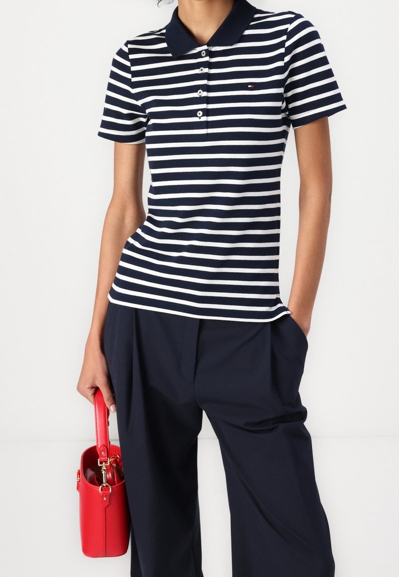 Personne portant un polo rayé bleu marine et blanc et un pantalon bleu marine, tenant un petit sac à main rouge avec des accessoires dorés dans une main.