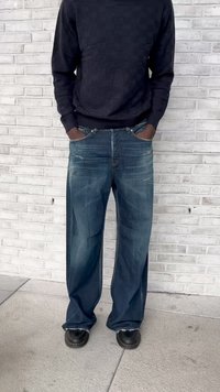 Blå denim jeans med en løs, vid bendesign. Kombinert med en mørk, strukturert genser, og svarte sko. Stående mot en murvegg.