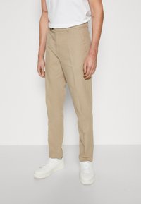 Pantalons en lin beiges avec une coupe droite, dotés d'une fermeture éclair, de poches latérales et d'une surface texturée. Associés à des baskets blanches.