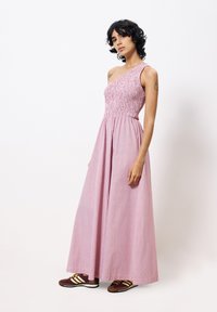 Robe maxi rose à une épaule avec un corsage smocké, un tissu fluide et un motif rayé subtil. Portée avec des chaussures de sport marron et jaune.