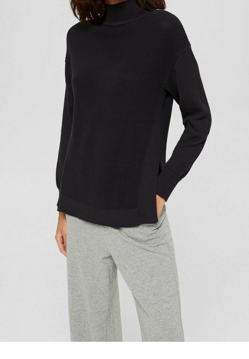 Maglione nero a maglia con colletto alto, spalle scese, fessure laterali e polsini a coste. Abbinato a pantaloni grigi chiari dalla vestibilità ampia.