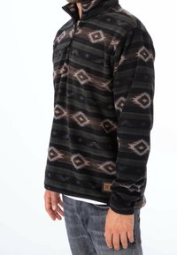 Svart fleece-pullover med en kvart dragkedja, som har ett geometriskt mönster i bruna och grå ränder. Texturerad tyg och logotypmärke vid fållen.