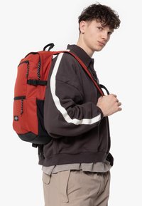 Sac à dos multi-poches rouge et noir avec des bretelles réglables, en tissu texturé et matériel noir, avec un logo blanc à l'avant.