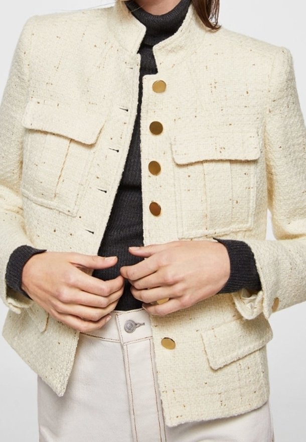 Femme portant une veste crème texturée boutonnée par-dessus un pull à col roulé noir, les mains ajustant la veste à la taille.