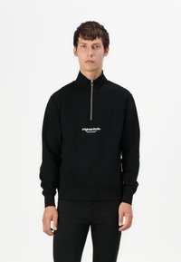 Crna zip-up majica s visokim ovratnikom. Ima bijeli natpis na prsima. Mekana tekstura i uski kroj. Nosena s crnim hlačama.