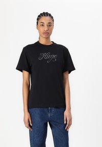 Black cotton t-shirt featuring a shiny "Hugo" logo in cursive, short sleeves, and a classic crew neckline. Paired with blue denim jeans.