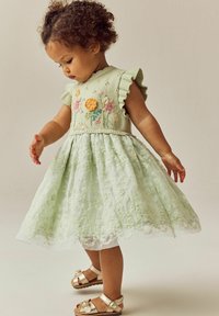 Lichtgroene jurk met geborduurde bloemenmotieven op het lijfje, doorschijnende kanten rok en flonkerende mouwen. Gouden sandalen maken de outfit compleet.