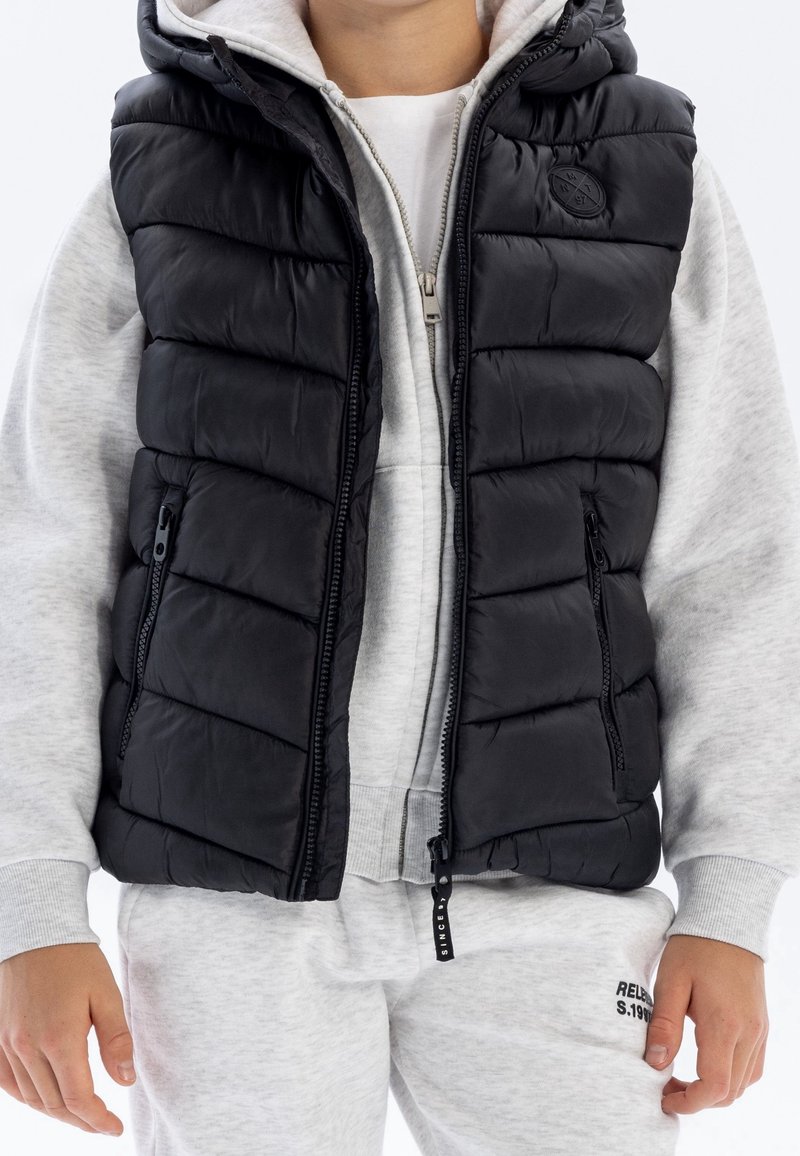 Enfant portant un sweat à capuche gris clair et un pantalon de survêtement, superposé avec une doudoune matelassée sans manches noire avec fermeture éclair à l'avant et des poches.