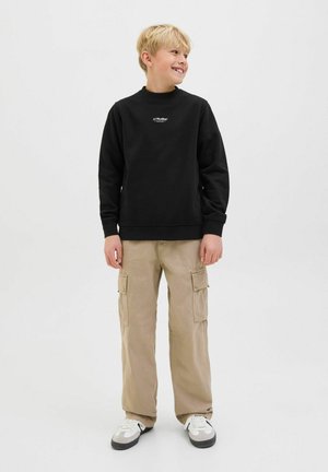 Fant s svetlimi lasmi, oblečen v črno jopico JJ Studios, bež hlače s številnimi žepi in bele superge, stoji in se smeji na enobarvnem ozadju.