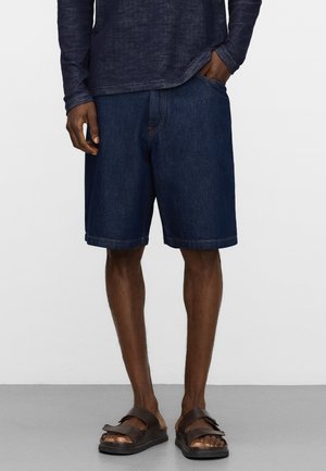 Man draagt donkerblauwe denim shorts, navy lange mouwen shirt en bruine sandalen, staat met één hand in zak op grijze vloer.
