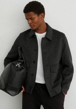 Uomo con giacca grigio scuro abbottonata e camicia bianca che tiene una grande borsa in pelle nera contro una parete chiara.