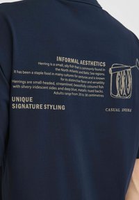 Tengerészkék polo ing, melynek hátulján nyomtatott szöveg és illusztrációk találhatók, a heringről és annak jellemzőiről világos betűtípussal.